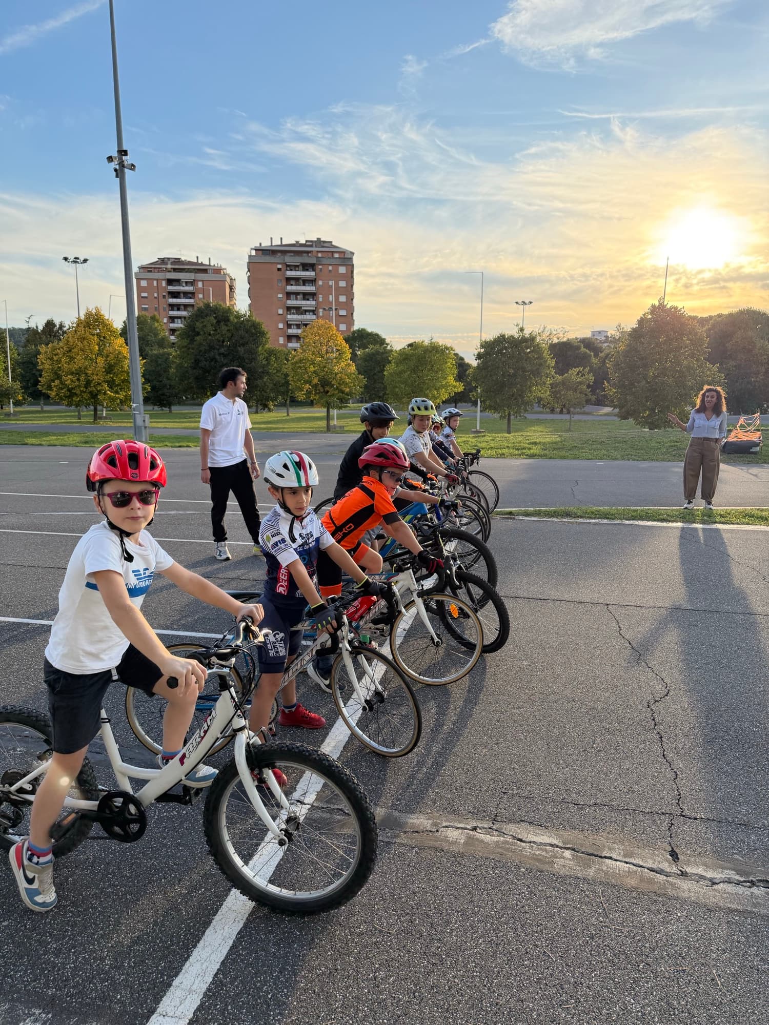 Fila di giovani allievi della Scuola Triono in sella alle bici al Ciclodromo Renato Perona, luce del tramonto, maestri accanto